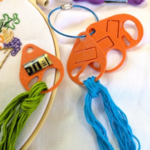 May include: A beige embroidery hoop containing fabric with floral stitching, accompanied by orange plastic thread organisers. One organiser holds green embroidery floss labelled 861, while others hold blue floss. A blue cable ring and purple floss are present.