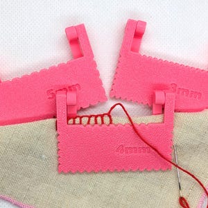 May include: Three bright pink plastic sewing spacers with serrated edges, labelled 3mm, 4mm, and 5mm. One spacer is attached to a piece of beige fabric with red thread and a metal needle, while the other two spacers are positioned above the fabric.