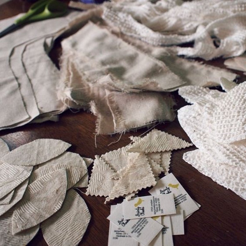 May include: A collection of fabric pieces in various shapes and textures, including leaf-shaped cutouts, torn strips, and crocheted sections. The color palette is primarily off-white and beige, with small tags featuring text and a yellow bird logo.