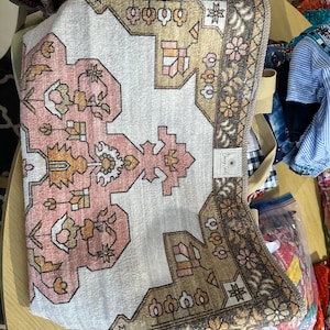 May include: A folded rectangular beach towel with a rug-like design. The towel features a white background with a pink and brown geometric pattern. The border is brown with floral accents. The towel has a beige strap.