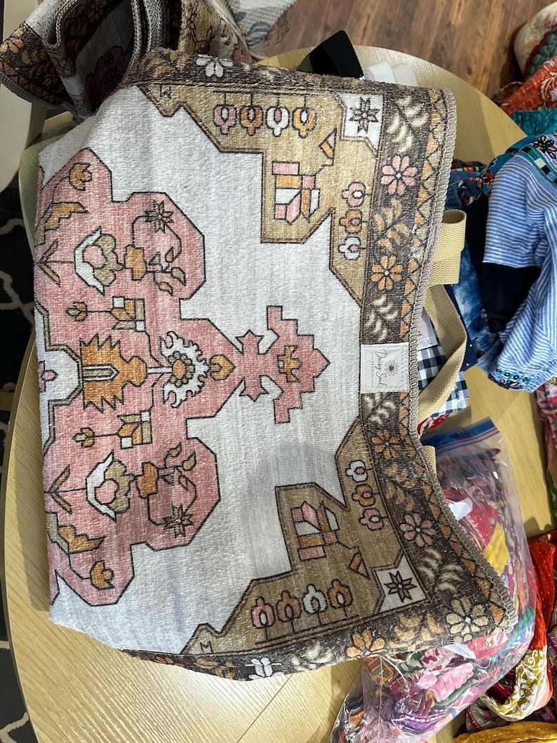 May include: A folded rectangular beach towel with a rug-like design. The towel features a white background with a pink and brown geometric pattern. The border is brown with floral accents. The towel has a beige strap.