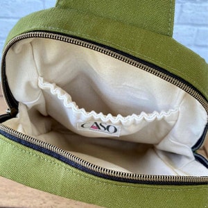 May include: Olive green canvas bag with a black zipper and a white interior. The bag has a small pocket inside and a tag that reads "CASO". The bag is open, showing the interior.