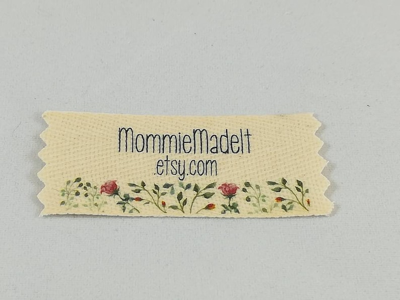 May include: A beige fabric label with a scalloped edge and a floral design. The label has the text "MommieMadeIt etsy.com" printed on it.