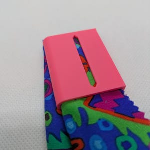 May include: A bright pink rectangular belt buckle attached to a fabric belt featuring a vibrant, multi-colored abstract pattern with shades of blue, green, purple, and orange. The buckle has a central vertical slot and a smooth, matte finish.