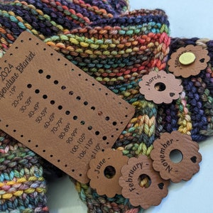 May include: A brown leather temperature blanket marker with holes for each month of the year. The marker is labeled with the text "2024 Temperature Blanket" and includes a temperature scale from -19 degrees to over 110 degrees Fahrenheit. The marker is placed on a colorful knitted blanket with different colored stripes.