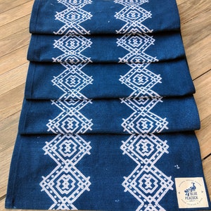 May include: Folded indigo blue table runner with a white geometric diamond pattern. The runner is made of fabric and has a tag that reads "Blue Peacock, Natural Indigo Goods, Hand Printed & Dyed."