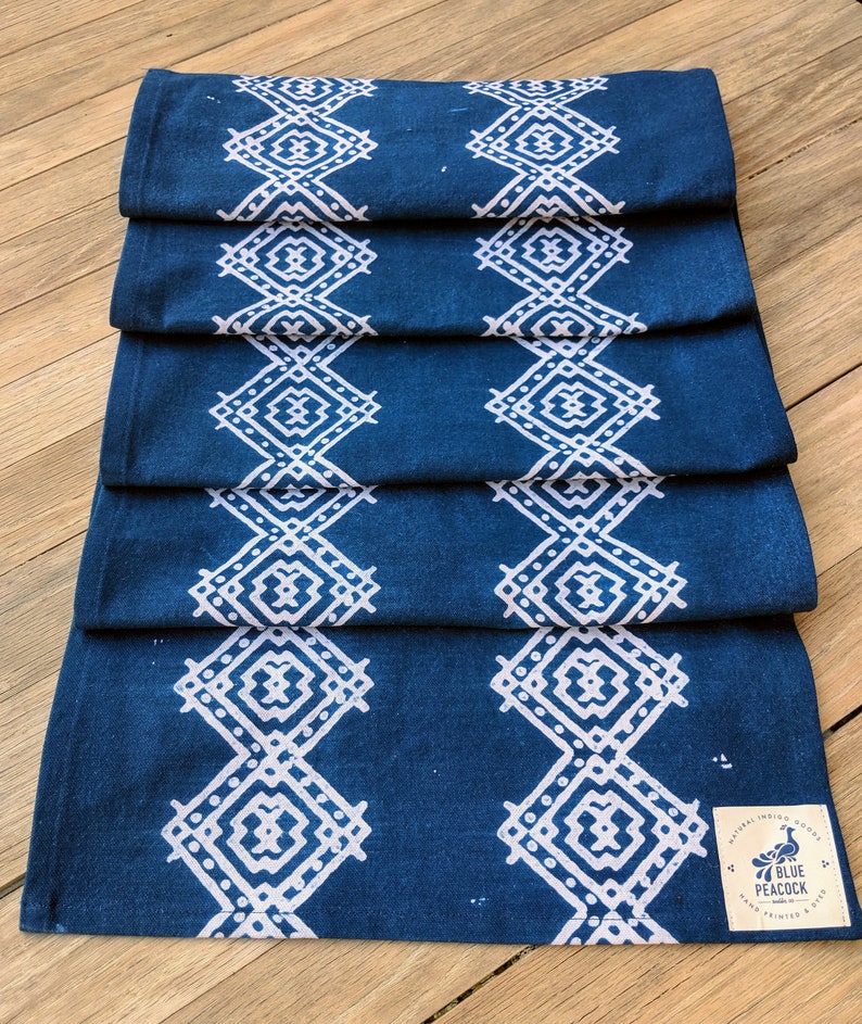 May include: Folded indigo blue table runner with a white geometric diamond pattern. The runner is made of fabric and has a tag that reads "Blue Peacock, Natural Indigo Goods, Hand Printed & Dyed."