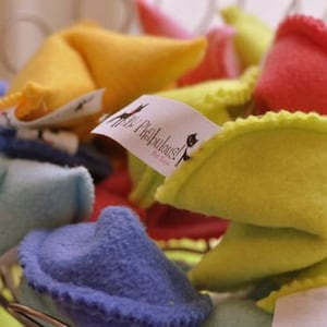May include: A collection of colorful, plush fortune cookies in shades of yellow, red, blue, and pink. Some cookies have tags with the text "Purr-Fabulous!" and a cat silhouette. The cookies are in a wire basket.