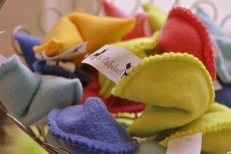 May include: A collection of colorful, plush fortune cookies in shades of yellow, red, blue, and pink. Some cookies have tags with the text "Purr-Fabulous!" and a cat silhouette. The cookies are in a wire basket.