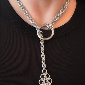 Puede incluir: Collar de cota de malla plateado con un gran colgante circular y un elemento colgante decorativo de cota de malla. El collar está hecho de anillos de metal entrelazados, creando un diseño texturizado y detallado. El collar se usa sobre un fondo negro.
