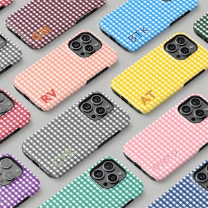 May include: A collection of phone cases in various colors, including pink, yellow, blue, and gray. Each case features a gingham pattern and a monogram of two or three letters. The cases are designed to fit a smartphone.