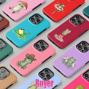 May include: A selection of brightly coloured phone cases, each adorned with a different frog illustration. Cases are in pink, blue, red, and purple. Designs include a frog in sunglasses, a frog in a cowboy hat, and a frog in a tutu.
