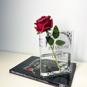 Könnte beinhalten: Eine transparente Acrylvase mit dem Text "The Phantom of the Opera" und einem dekorativen Rand, die eine rote Rose enthält. Die Vase steht auf einem Buch mit dem Titel "The Complete Phantom of the Opera".