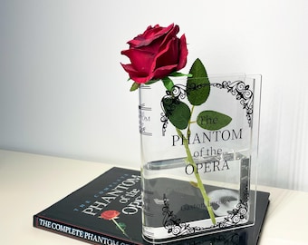 May include: A clear acrylic vase with the text "The Phantom of the Opera" and a decorative border, holding a red rose. The vase sits atop a book titled "The Complete Phantom of the Opera".