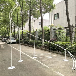 Puede incluir: Una estructura de arco de metal blanco con una parte superior curva y soportes verticales. El arco está instalado al aire libre sobre una superficie pavimentada, con árboles y edificios al fondo. La estructura está diseñada para eventos.