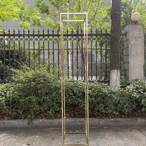 Puede incluir: Un marco rectangular alto de metal dorado, probablemente para fines decorativos. El marco está hecho de tubos metálicos cuadrados y delgados y se alza sobre una superficie de hormigón. El fondo incluye vegetación, una valla de hierro forjado y un edificio.