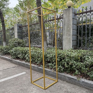 Könnte beinhalten: Ein hoher, rechteckiger Metallrahmen in Gold, wahrscheinlich für dekorative Zwecke. Der Rahmen besteht aus Vierkantrohren und steht auf einer Betonoberfläche. Der Rahmen befindet sich im Freien, mit Grün und einem Zaun im Hintergrund.