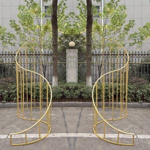 以下が含まれることがあります： 屋外イベントや庭園用の、2つの金色の湾曲した金属構造。構造はらせん状のデザインで、木、フェンス、建物を背景に配置されています。