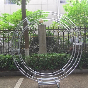 Könnte beinhalten: Eine silberne, kreisförmige Bogenstruktur aus Metall mit drei konzentrischen Ringen, die von einer rechteckigen Basis getragen wird. Der Bogen befindet sich im Freien, vor dem Hintergrund von grünem Laub, einem Baum und einem Gebäude.