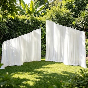 Può includere: Due fondali in tessuto bianco autoportanti con drappeggi raccolti e plissettati. Le strutture presentano un design a gradini e angolato con materiale morbido e fluido che si estende fino a terra, creando un aspetto pulito e stratificato.