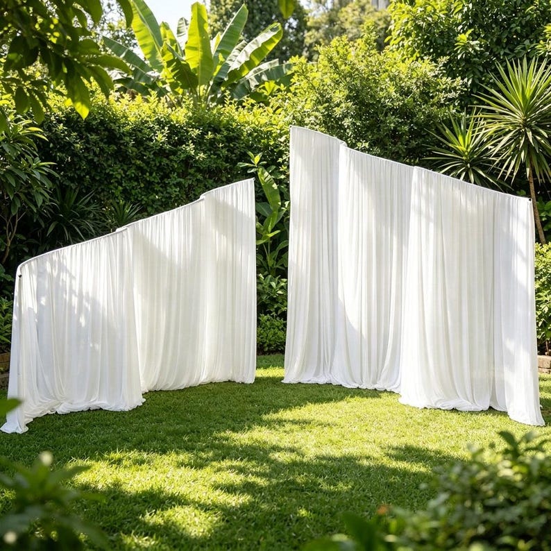 May include: Two freestanding white fabric backdrops with gathered, pleated drapes. The structures feature a tiered, angled design with soft, flowing material that extends to the ground, creating a clean, layered appearance for outdoor settings.