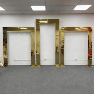 Könnte beinhalten: Drei goldfarbene, rechteckige Torbogenstrukturen. Die Bögen bestehen aus einem reflektierenden Material, wahrscheinlich Metall, und sind in einer Reihe angeordnet. Die Strukturen stehen auf einem grauen Boden vor einer weißen Wand.