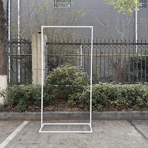 May include: A white metal rectangular frame stands outdoors. The frame is open in the centre and has a base. The background includes greenery, a fence, and a concrete wall.