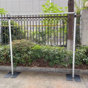 May include: A silver telescoping backdrop stand with black bases. The stand is set up outdoors against a green bush and a black metal fence.