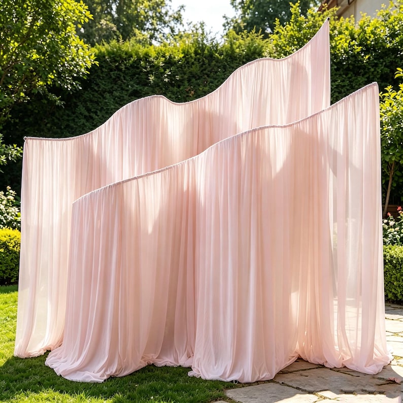 May include: A pale pink fabric backdrop featuring a series of tiered, wave-shaped frames draped with sheer, pleated material. The structure consists of multiple overlapping panels that create a layered, flowing appearance with soft vertical folds.