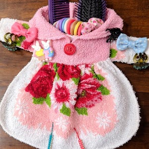 May include: A pink and white towel shaped like a dress, decorated with red roses and hair accessories. The dress has a pink hood and is adorned with hair ties, combs, and decorative bows. Two small bunny figurines are attached to the dress.