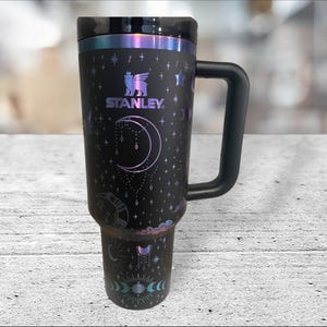 May include: A black stainless steel travel mug with a black handle. The mug has a celestial design with a crescent moon, stars, and clouds. The mug has the Stanley logo and the text "Stanley" on it.