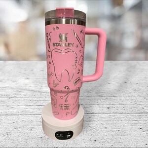 May include: Pink Stanley travel mug with a handle. The mug is decorated with a large tooth and dental tools in a gold metallic colour. The text "Stanley" is printed on the mug.
