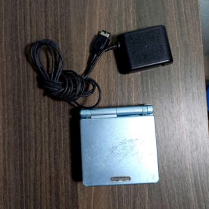 Op de afbeelding: Een lichtblauwe Nintendo Game Boy Advance SP handheld gameconsole met een zwarte voedingsadapter en snoer. De console is open en toont het Nintendo-logo. De voedingsadapter is zwart.