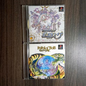 Op de afbeelding: Twee PlayStation-gamecases. De bovenste case heeft een kleurrijke illustratie van personages met de titel "PoPoLoCrois" in het Engels en Japans. De onderste case heeft een vergelijkbaar ontwerp met de titel "PoPoLoCrois".