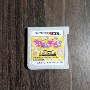 May include: A Nintendo 3DS game cartridge with a colorful label featuring Japanese text and illustrations of pastries. The label includes the text "Alchemist" and the Nintendo 3DS logo.