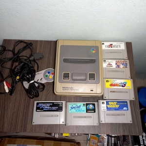May include: A beige Nintendo Super Famicom console with a gray controller and several game cartridges. The cartridges include titles like "Final Fantasy VI" and "Space Invaders". The console and accessories are on a wooden surface.