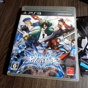 Puede incluir: Caja de juego de PS3 para "BlazBlue: Continuum Shift". La carátula presenta múltiples personajes de estilo anime en poses dinámicas, con una combinación de colores azul y blanco. La caja del juego está en una funda de plástico transparente.