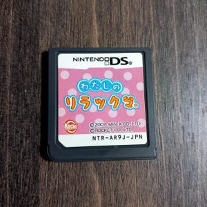 May include: A Nintendo DS game cartridge with a pink label featuring the text "わたしのリラックマ" in Japanese. The label also includes the Nintendo DS logo, the Rocket logo, and copyright information. The cartridge is gray and sits on a wooden surface.