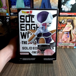 Może przedstawiać: Pudełko z figurką Dragon Ball Z Solid Edge Works z Friezą. Pudełko jest czarne z białym tekstem i zdjęciem figurki Friezy. Figurka jest fioletowa, biała i brązowa.