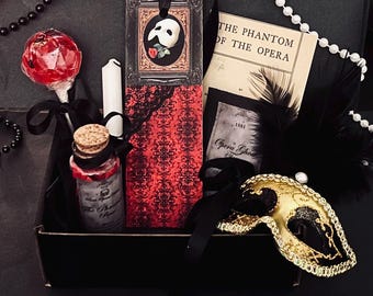 May include: A black gift box containing a red and black patterned bookmark with a Phantom mask image, a gold and black masquerade mask, a glass bottle with a cork stopper, a red lollipop, a white candle, and a book titled The Phantom of the Opera.