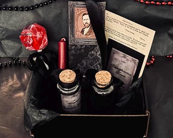 May include: A black gift box containing two glass bottles with cork stoppers, a red lollipop, a red candle, a portrait card, and a printed page from Dracula. The items are arranged with black ribbon and black tissue paper, accented by a string of black and red beads.