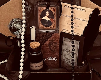 May include: A gift box containing a bookmark with a portrait of Mary Shelley, a small glass bottle with a cork stopper labeled Dr. Frankenstein, a black feather, a lollipop, a string of white pearls, a string of black beads, and a card titled Frankenstein.
