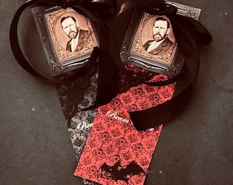 May include: Two rectangular bookmarks featuring a sepia-toned portrait of a man in a vintage frame, topped with a black satin ribbon bow. One bookmark has a red background with a black damask pattern and bat silhouette, the other is grey.