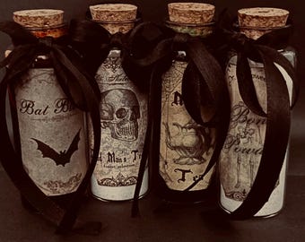Può includere: Quattro bottiglie di vetro trasparente con tappi di sughero, ciascuna legata con un nastro nero. Le etichette presentano grafiche in stile vintage e testo che include Bat Blood, Dead Man's Toe, Newt Tea e Bone Powder, con illustrazioni di pipistrello e teschio.