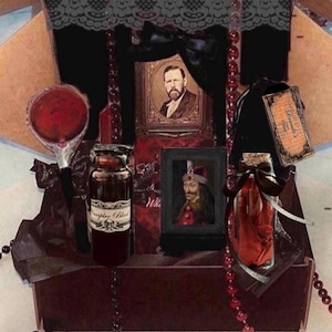 May include: A gift box containing a round red mirror, a glass bottle labeled Vampire Blood, a framed portrait of a man, a smaller framed portrait of a figure in a hat, a glass bottle with red fabric, and a tag labeled Dracula's Tea, accented with black lace and red beads.