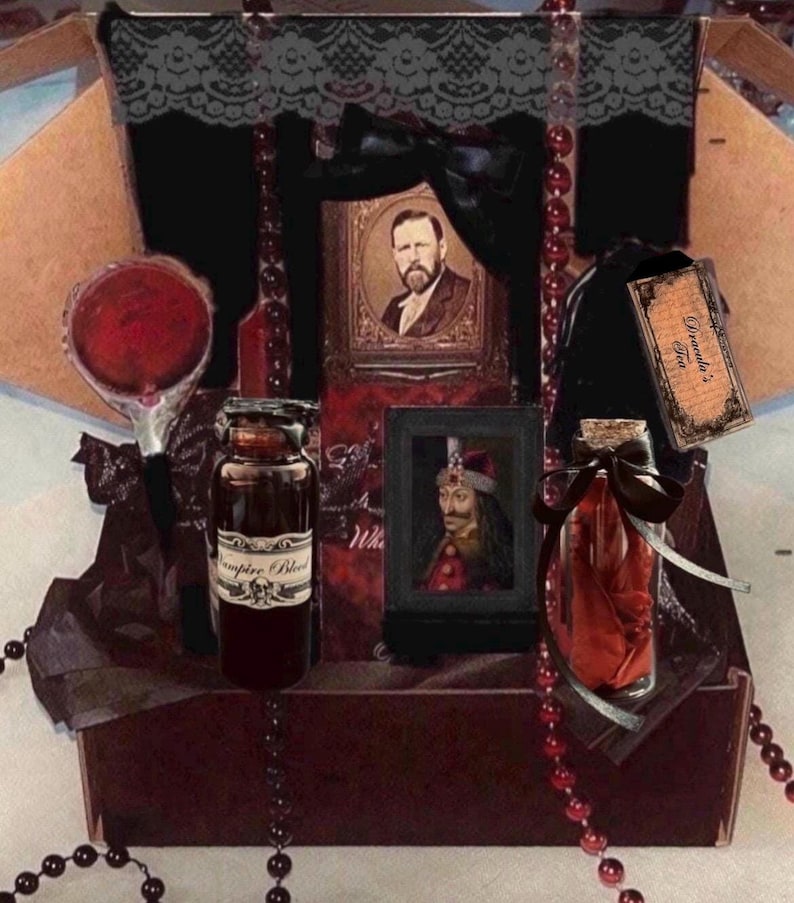 May include: A gift box containing a round red mirror, a glass bottle labeled Vampire Blood, a framed portrait of a man, a smaller framed portrait of a figure in a hat, a glass bottle with red fabric, and a tag labeled Dracula's Tea, accented with black lace and red beads.
