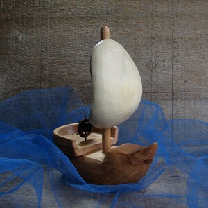 Può includere: Una barca a vela giocattolo di legno con una vela bianca, seduta su un tessuto di tulle blu. La barca a vela ha una piccola ruota di legno e un albero di legno.