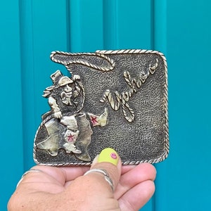 May include: A silver belt buckle featuring a cartoon cowboy riding a horse shaped like the state of Texas. The buckle says "Texas" in a stylized font.