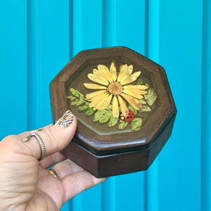 May include: A wooden octagonal box with a clear resin top featuring a pressed yellow daisy with a red ladybug. The box is brown and has a vintage aesthetic.