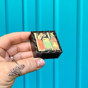 May include: A small, black decorative box with a hinged lid. The box features a colorful illustration of figures in period clothing. The box is held in a hand against a bright blue background.